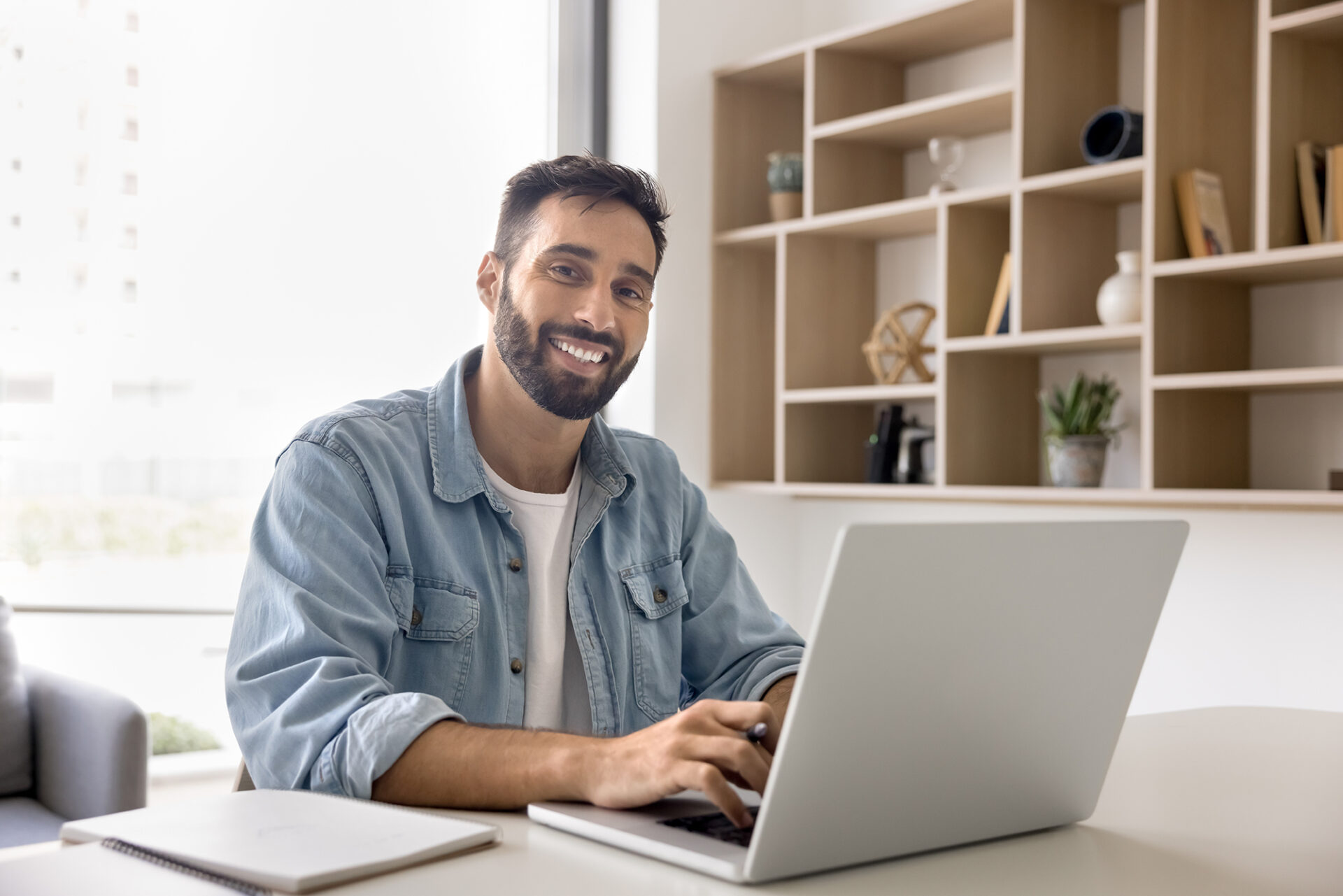 Happy man typing on laptop at office work table, looking at camera 